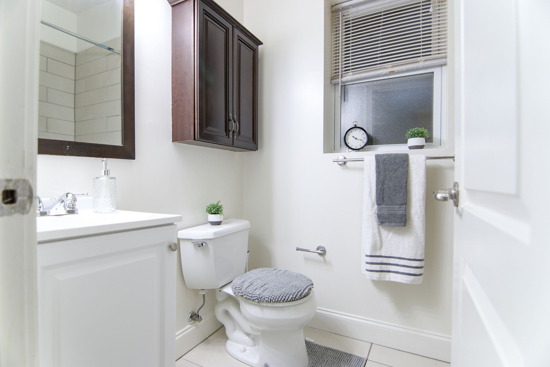 Small, white bathroom with a toilet, sink, and cabinet. A window with a towel rack and clock is visible.