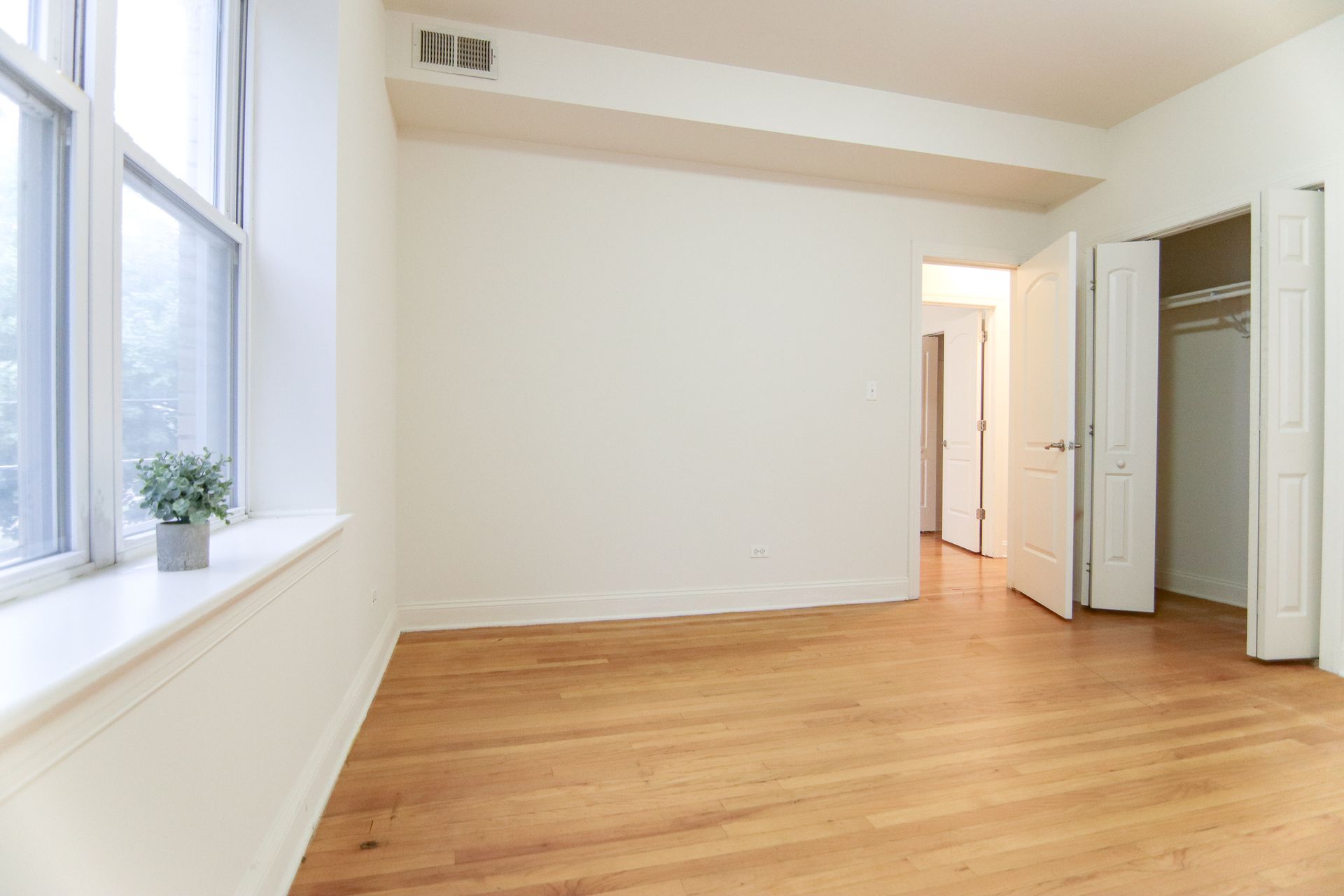 Empty bedroom with wooden floor, large window, open closet, and doorway.