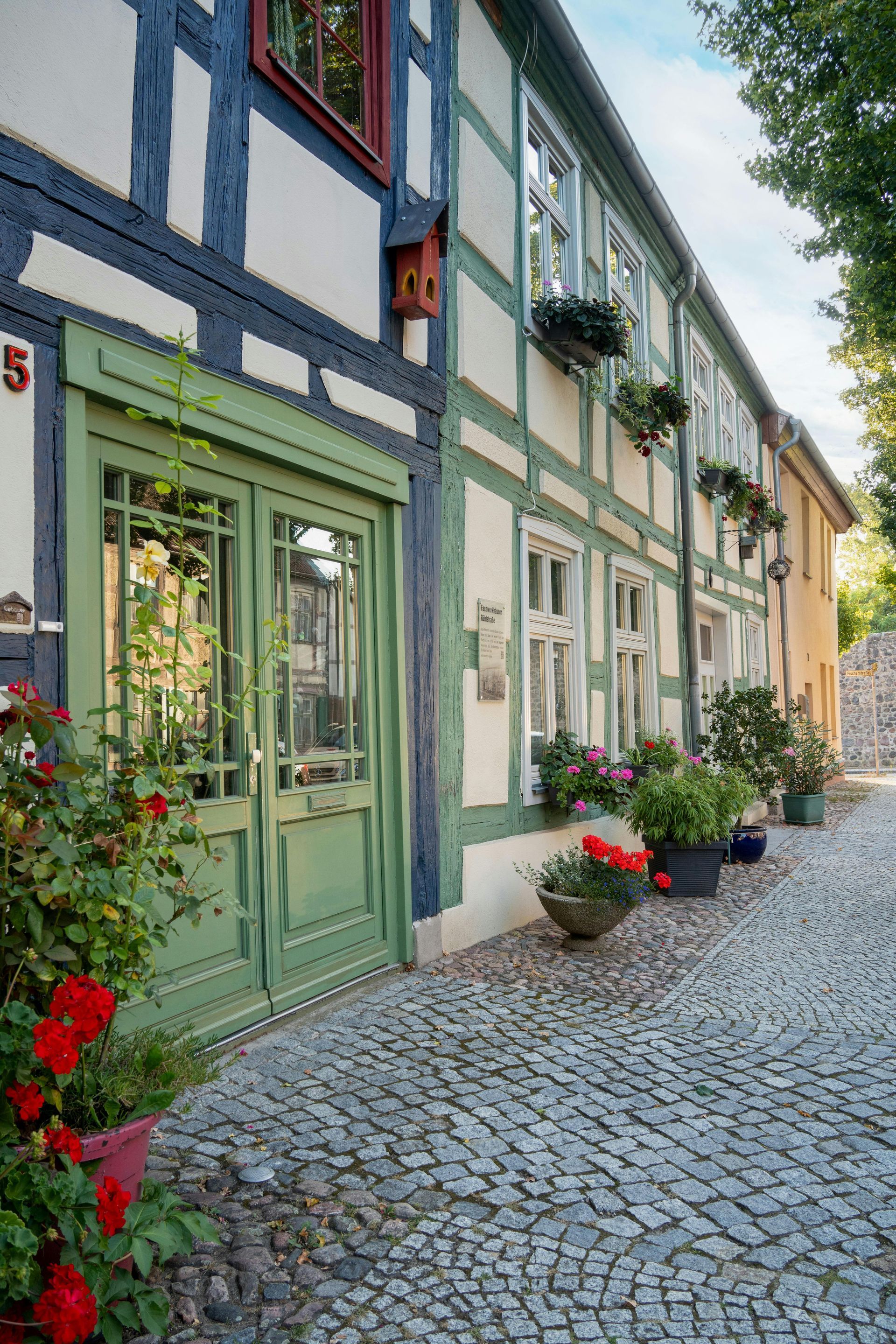 Vakwerkgebouw met groene deur en ramen, een straat met kinderkopjes en potplanten.