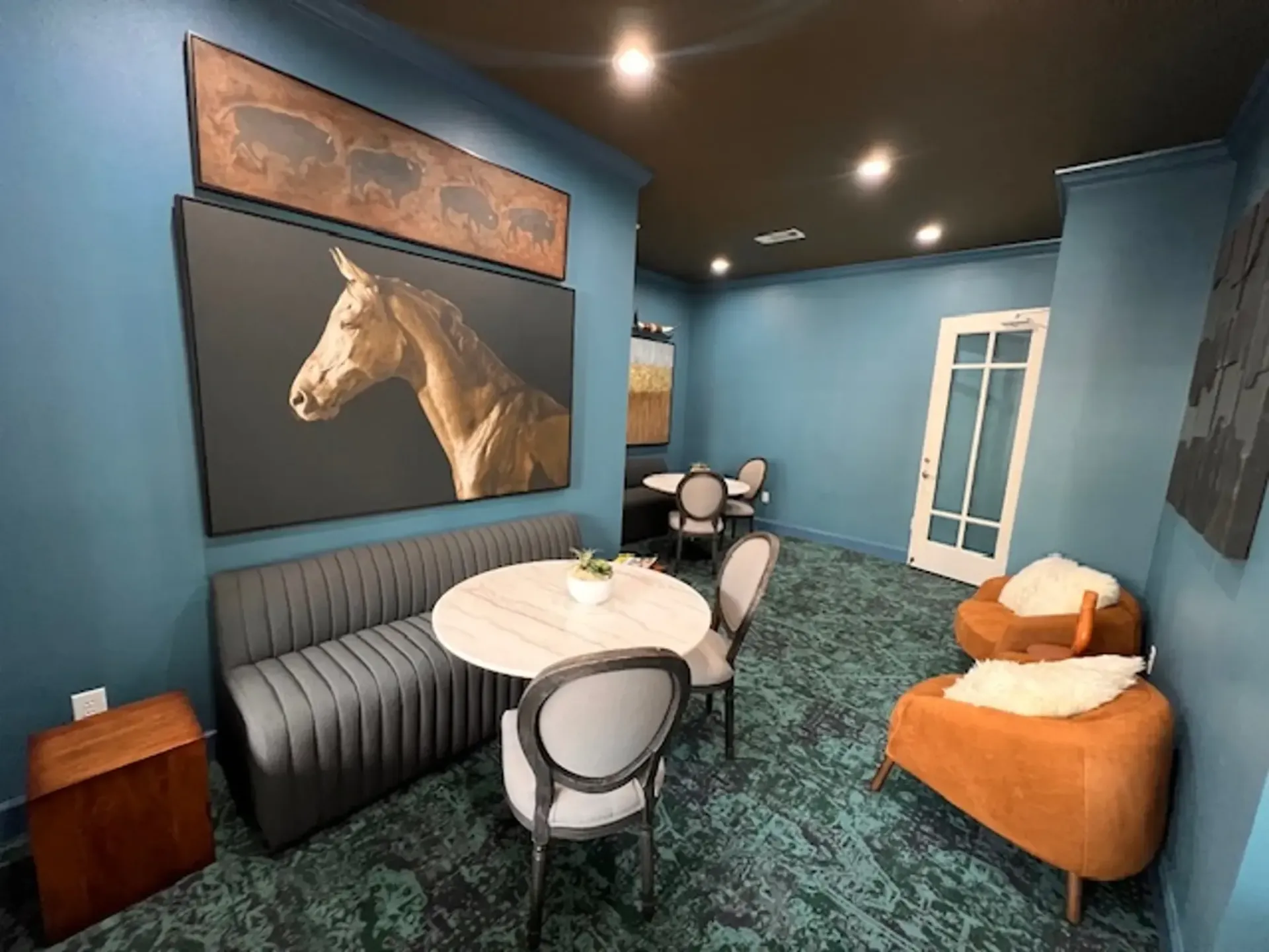 Resident clubhouse at Westpark Ranch Apartments in Houston, TX. A room with horse art, a round table with chairs, a sofa, and an orange armchair. Teal walls and green carpet.