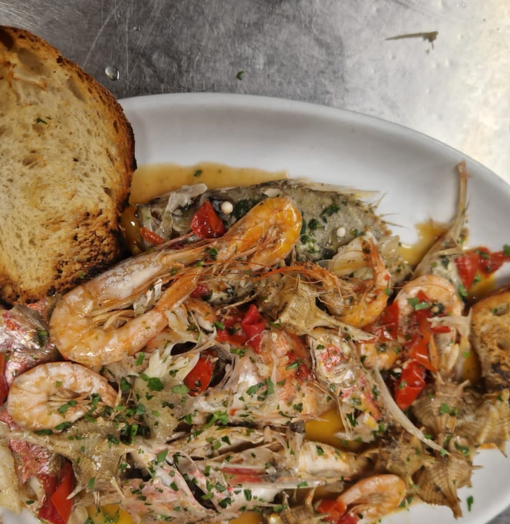 Stufato di pesce con gamberi, pesce, peperoni rossi, erbe aromatiche e pane tostato su un piatto bianco.