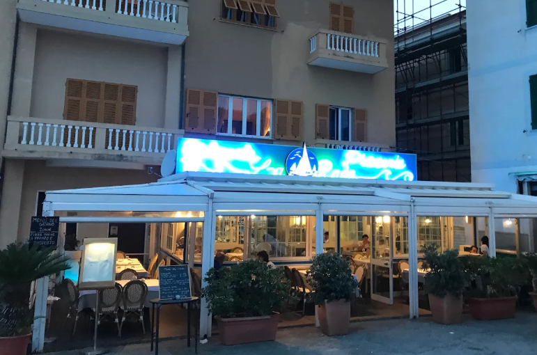 Esterno del ristorante con insegna illuminata blu, tenda da sole bianca, posti a sedere all'aperto, edificio sullo sfondo.