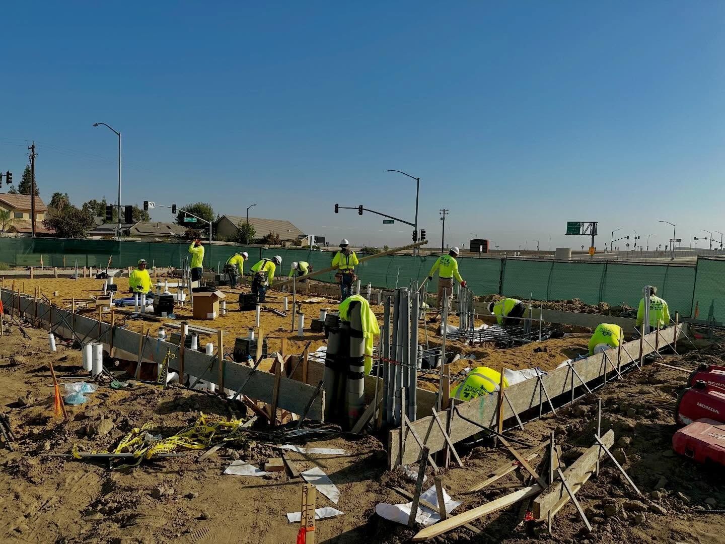 Construction workers in neon vests building concrete foundations at a roadside site under a clear blue sky