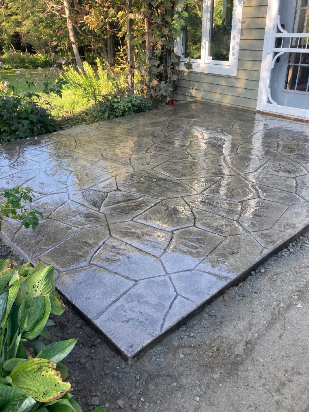 Wet stamped concrete patio beside a stone house, reflecting light after rain.