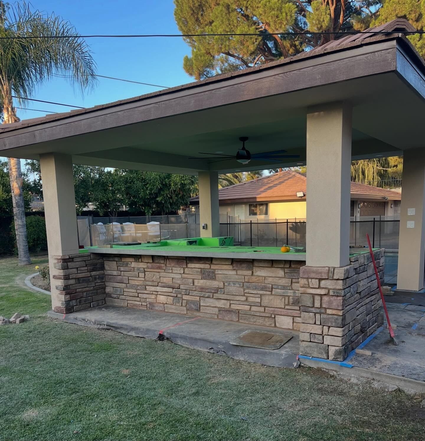Stone outdoor pavilion with a shingled roof and built-in bar, set in a grassy backyard.