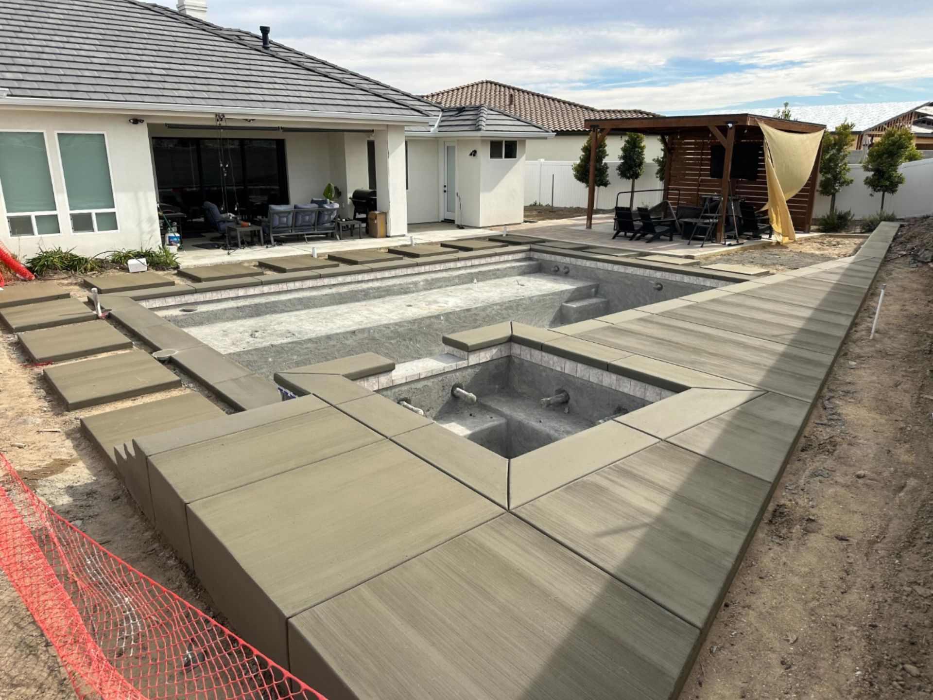 Backyard patio with a recessed pool and square hot tub under construction, bordered by tan decking.