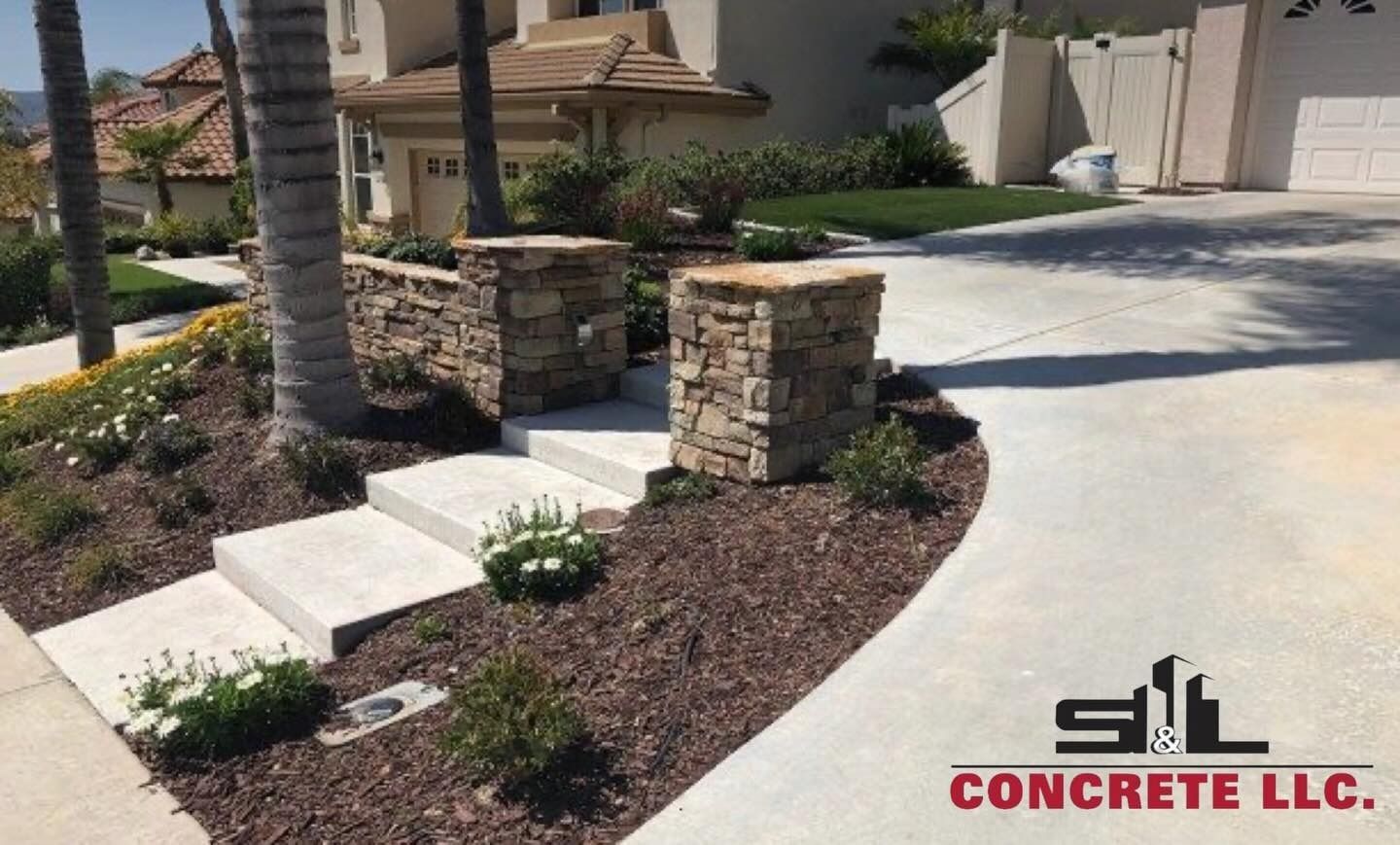 Concrete driveway and front walkway with stone pillar accents beside a house and landscaping.