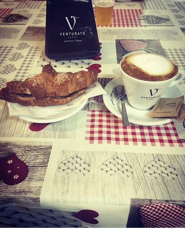 Croissant e cappuccino su un tavolo con un sacchetto di chicchi di caffè. Tovaglia a quadri rossi e bianchi.