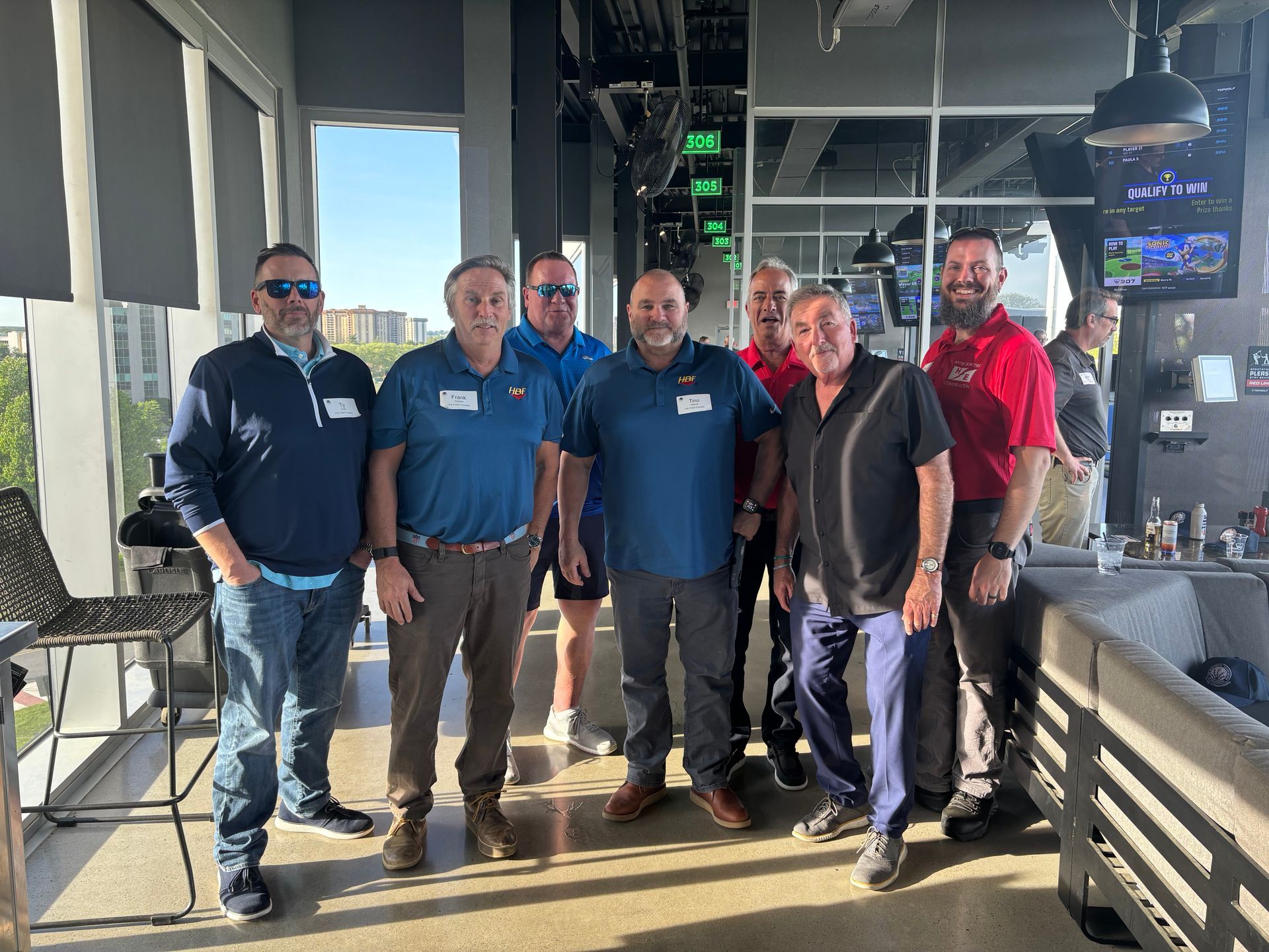 Six people at a Topgolf venue, posing for a photo. They stand near a window, smiling.