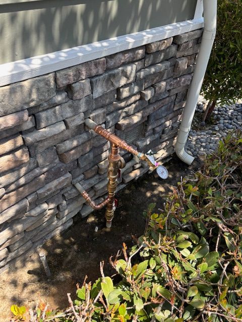 Copper pipes and valve next to a house with stone siding and gutter, near a bush.