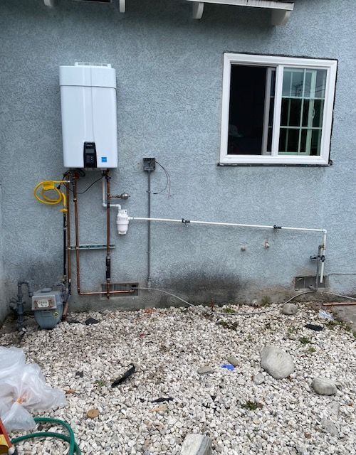 Exterior view of a wall-mounted water heater with gas and water lines. A window and pebble-covered ground are also visible.