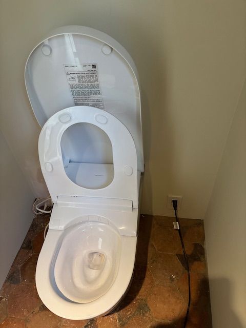 White bidet toilet with open seat, installed in a small, unfinished bathroom with a beige wall.