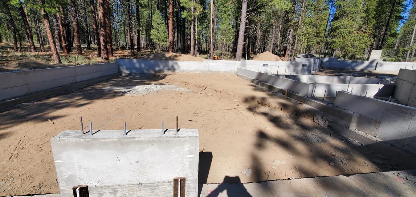 Construction site: concrete foundation with dirt fill, surrounded by trees.