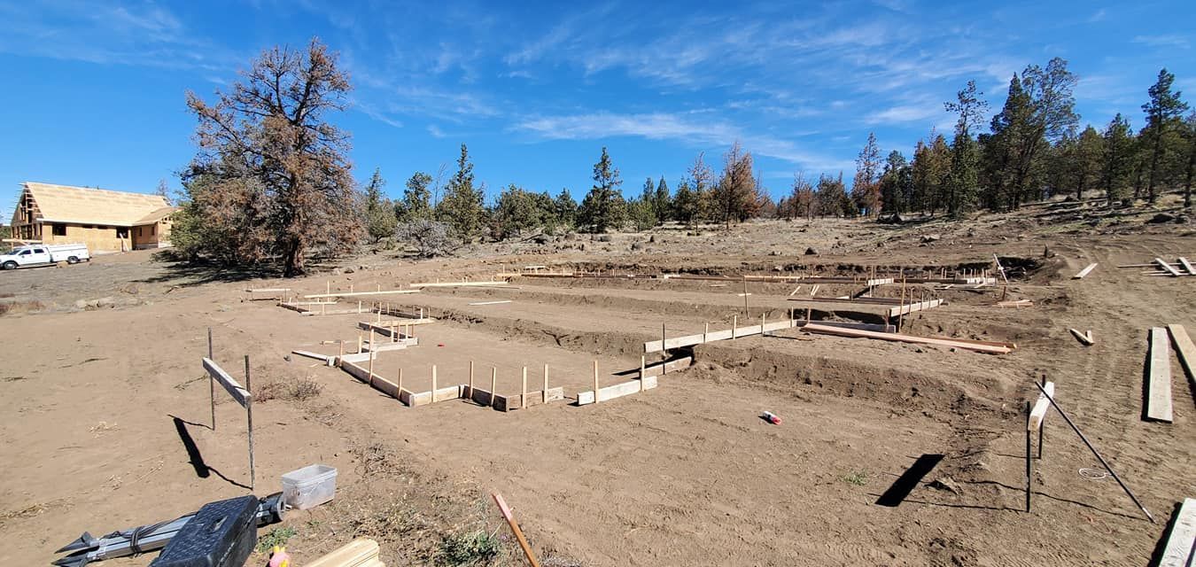 Construction site with foundation framework. Dirt, trees, and blue sky visible.
