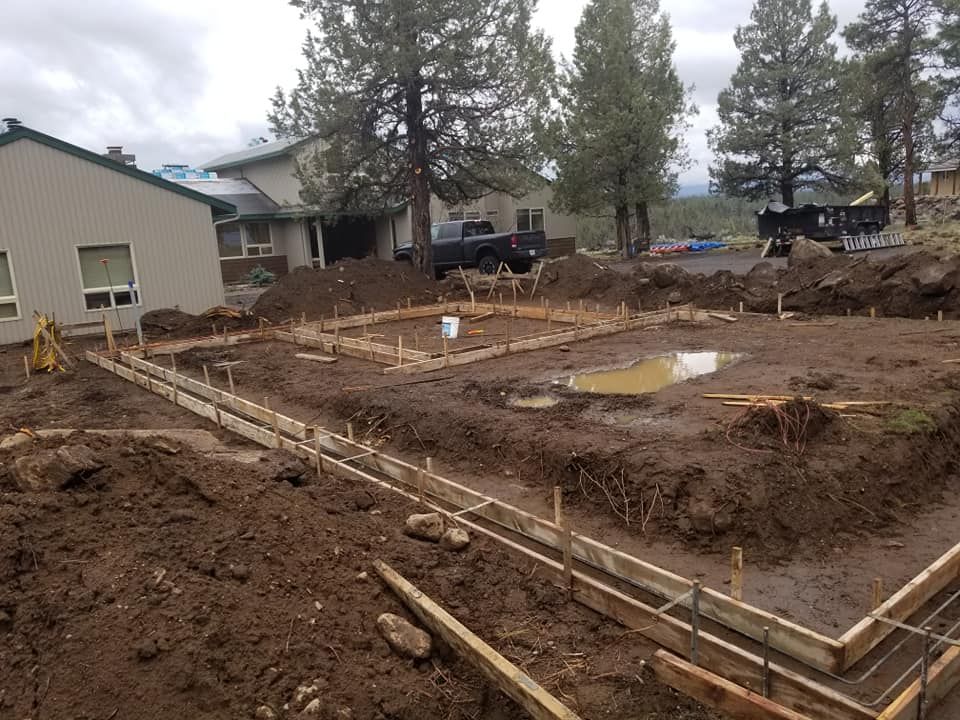 Construction site with wooden frames, dirt, and a partially built foundation. Houses and trees in the background.