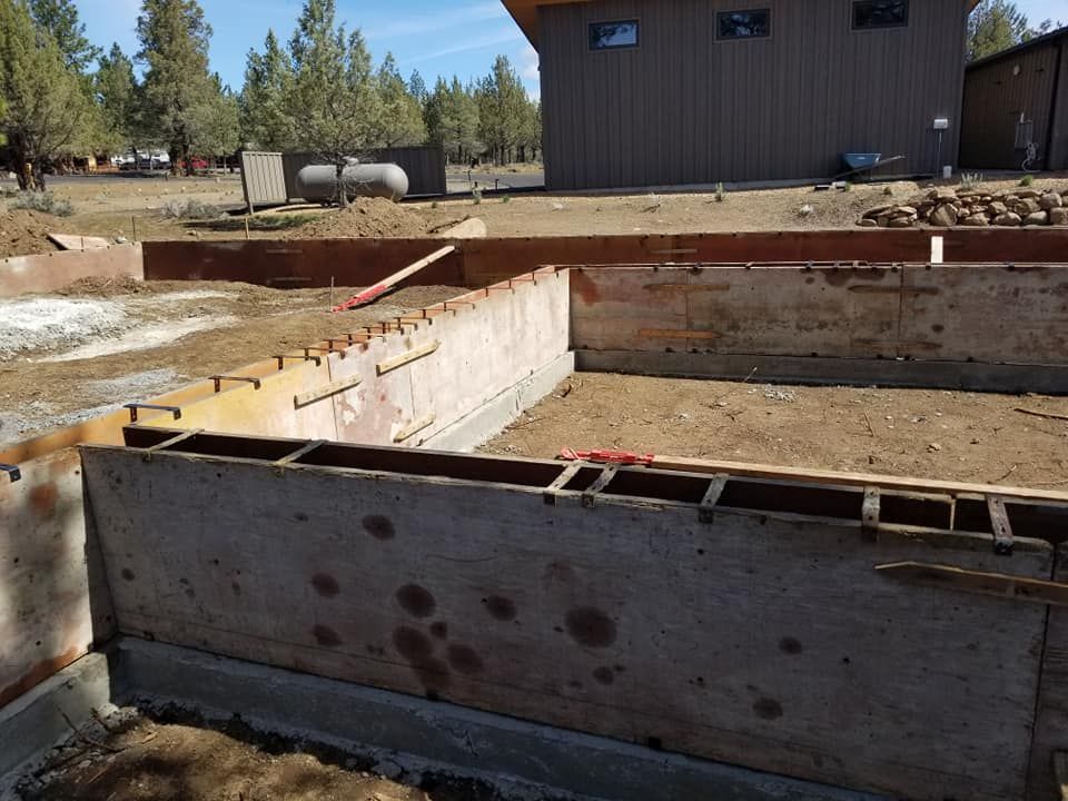 Wooden forms set for a foundation pour at a construction site, with a building in the background.
