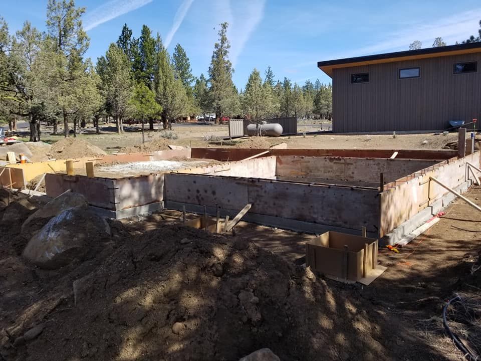 Construction site: Concrete foundation with wood forms, dirt, large rock, and a tan building in the background.