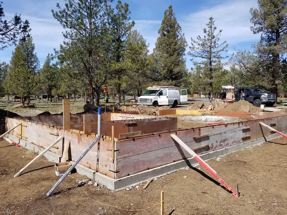 Foundation of a building under construction, with wooden forms and support beams, set in a wooded area.