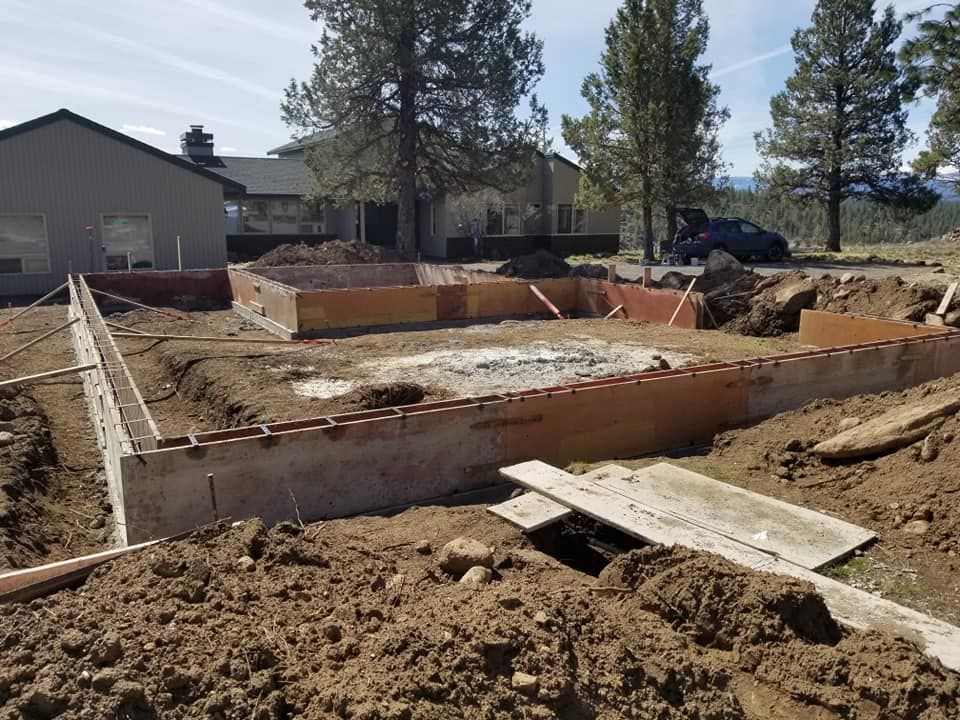 Construction site: Wooden forms outline a foundation. Brown earth surrounds the forms. A house and trees are in the background.