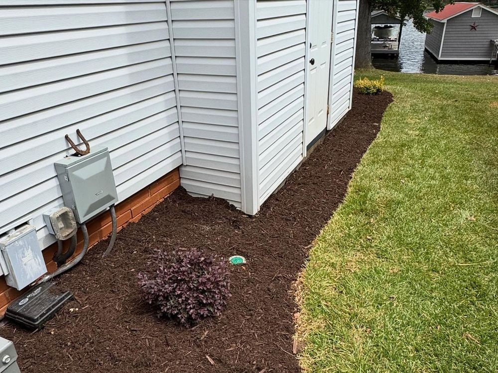 A building with white siding next to a mulched bed with a bush, alongside green grass and a body of water.