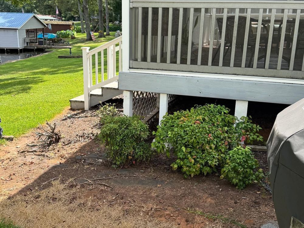 Garden bed with green bushes, under a gray deck with white railing, next to grass.