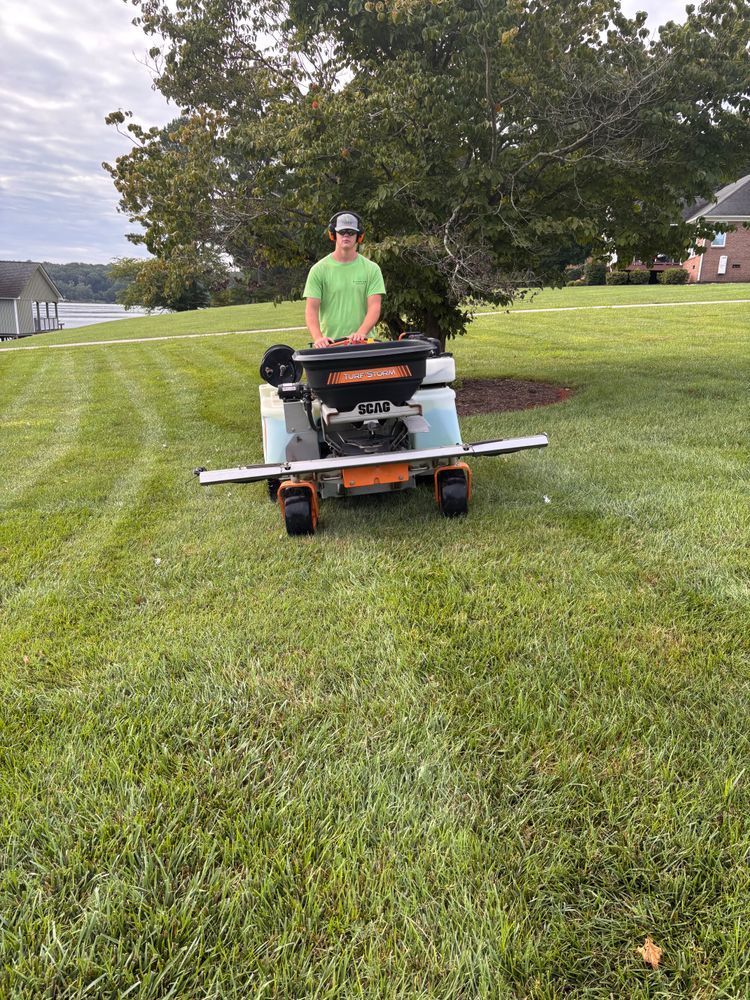 Person in a green shirt operating a spreader on a grassy lawn near water.