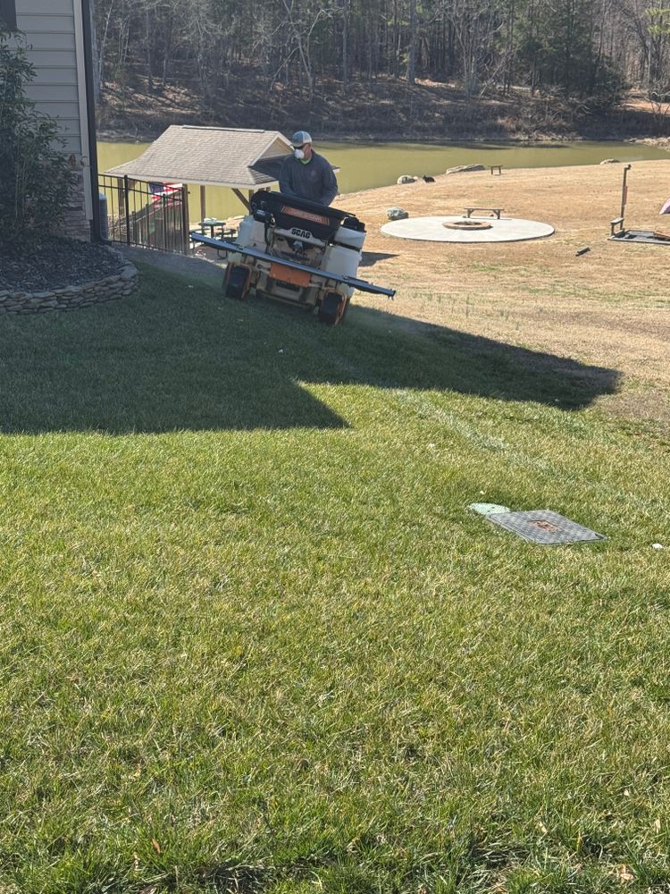 Person operating a yard machine on a grassy lawn near a pond.