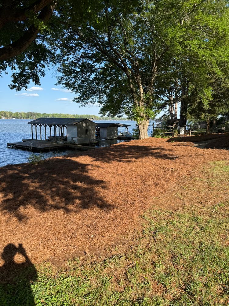 Lakeside dock with covered boathouse, large trees, and pine straw groundcover. Sunny day with water and blue sky.