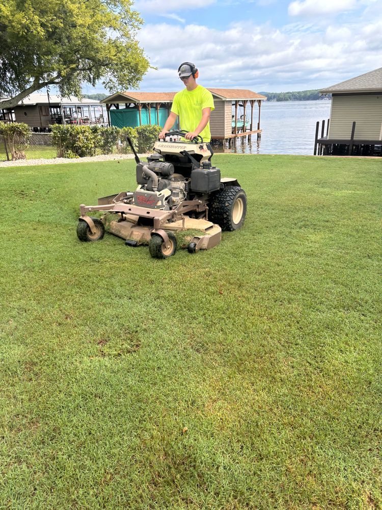 Person mowing a green lawn near a lake with a riding mower, wearing safety glasses and a neon yellow shirt.