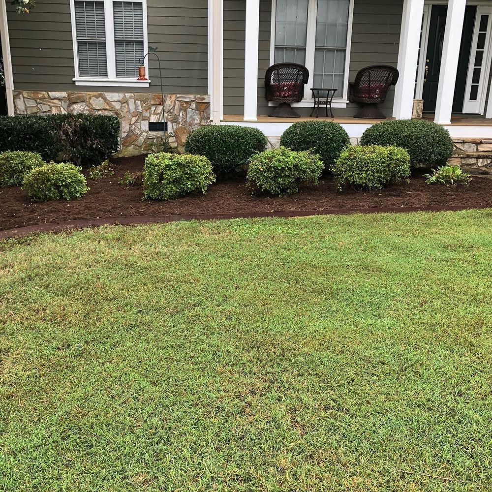 Green lawn and landscaped flower bed in front of a house with shrubs, mulch, and a porch.