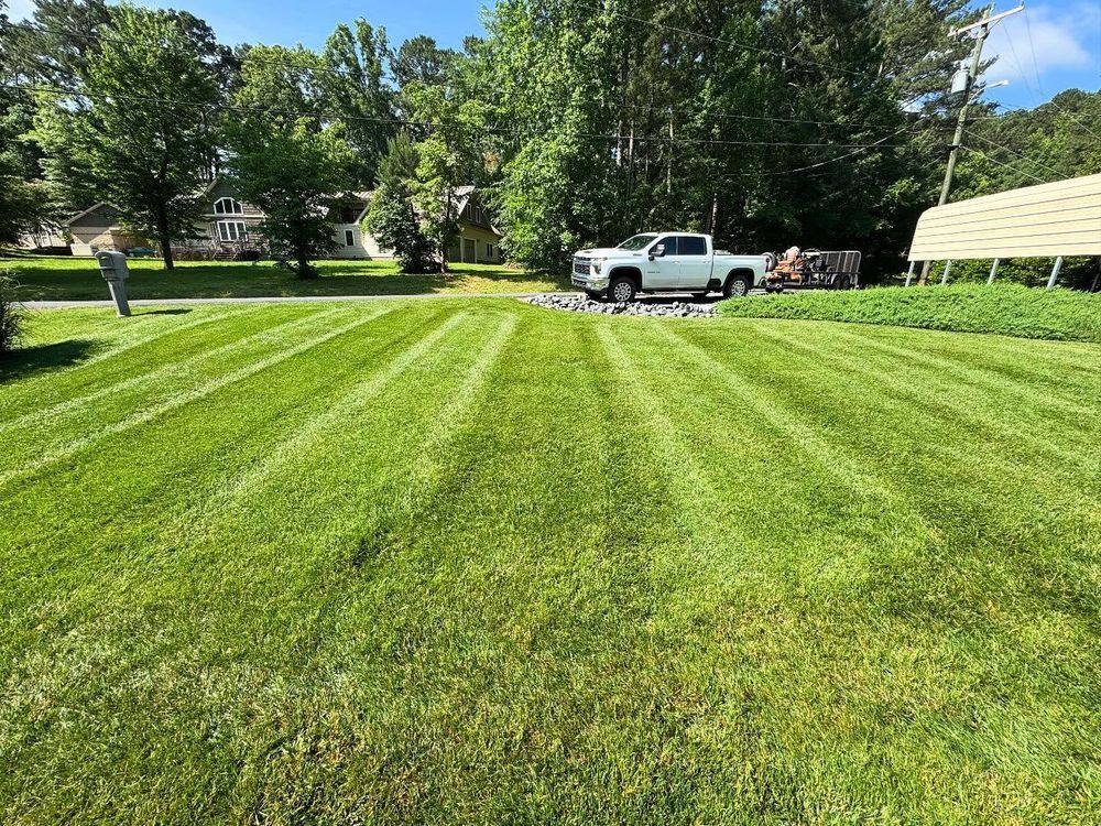 Lawn with freshly cut stripes; white pickup truck parked on gravel. Person standing on left, trees and sky in background.