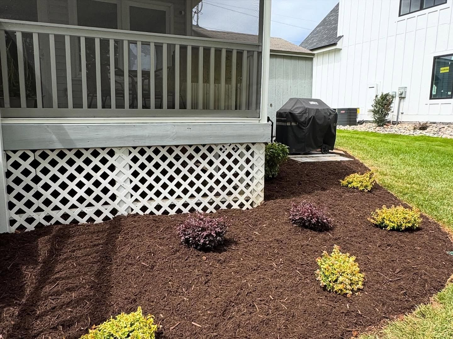 A backyard garden bed with dark mulch and small bushes under a porch with a grill.