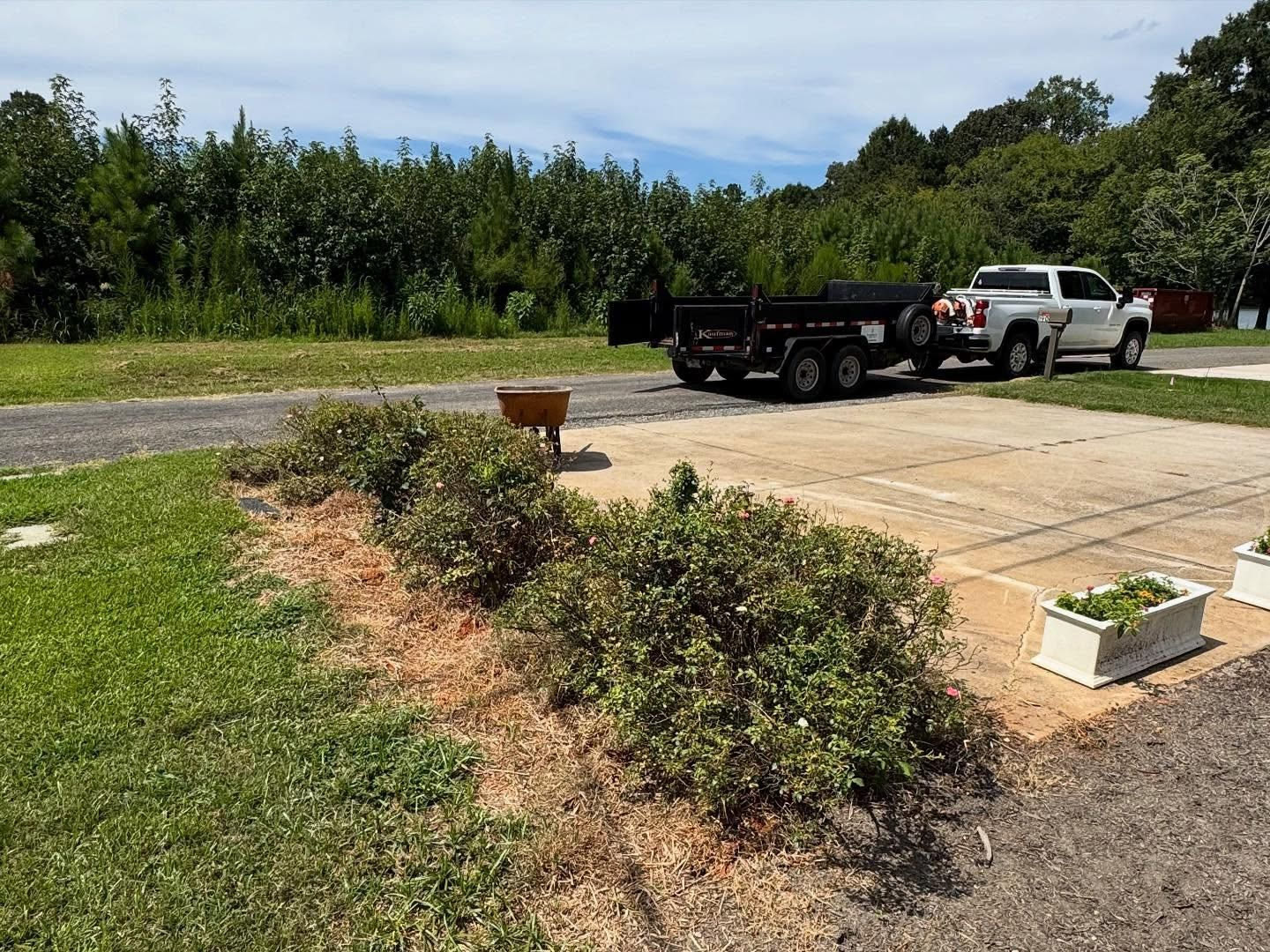 A white pickup truck with a trailer parked on concrete next to a landscaped area and a gravel path.