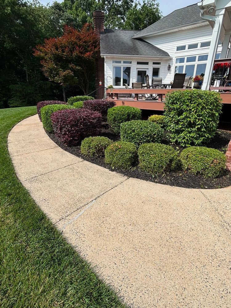 Concrete walkway curves past a landscaped garden with green and burgundy bushes, next to a house with a wooden deck.