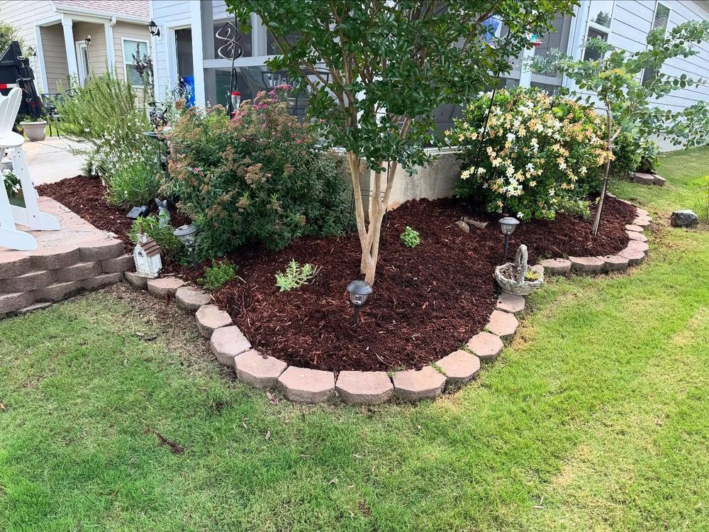 Landscaped garden bed with mulch, brick border, and various green plants around a tree.