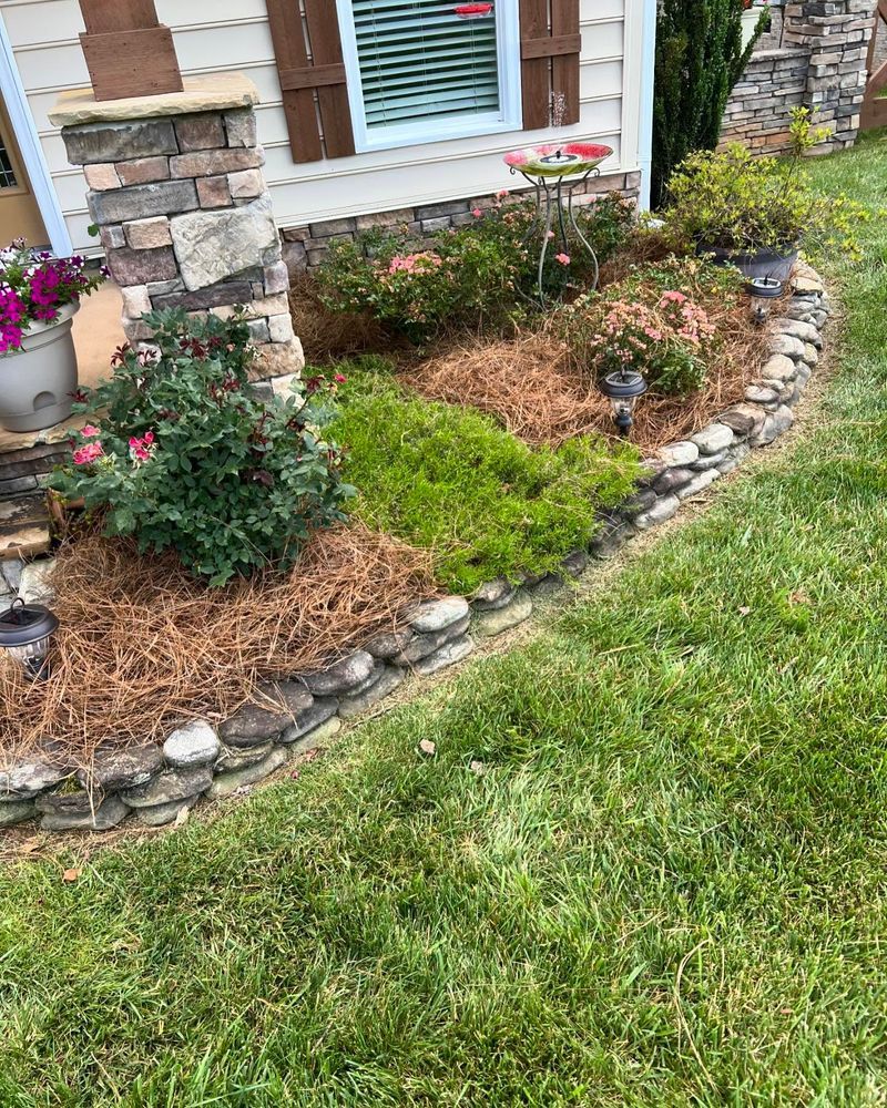 Flower bed with various plants and brown mulch, edged with gray stones, beside a house.