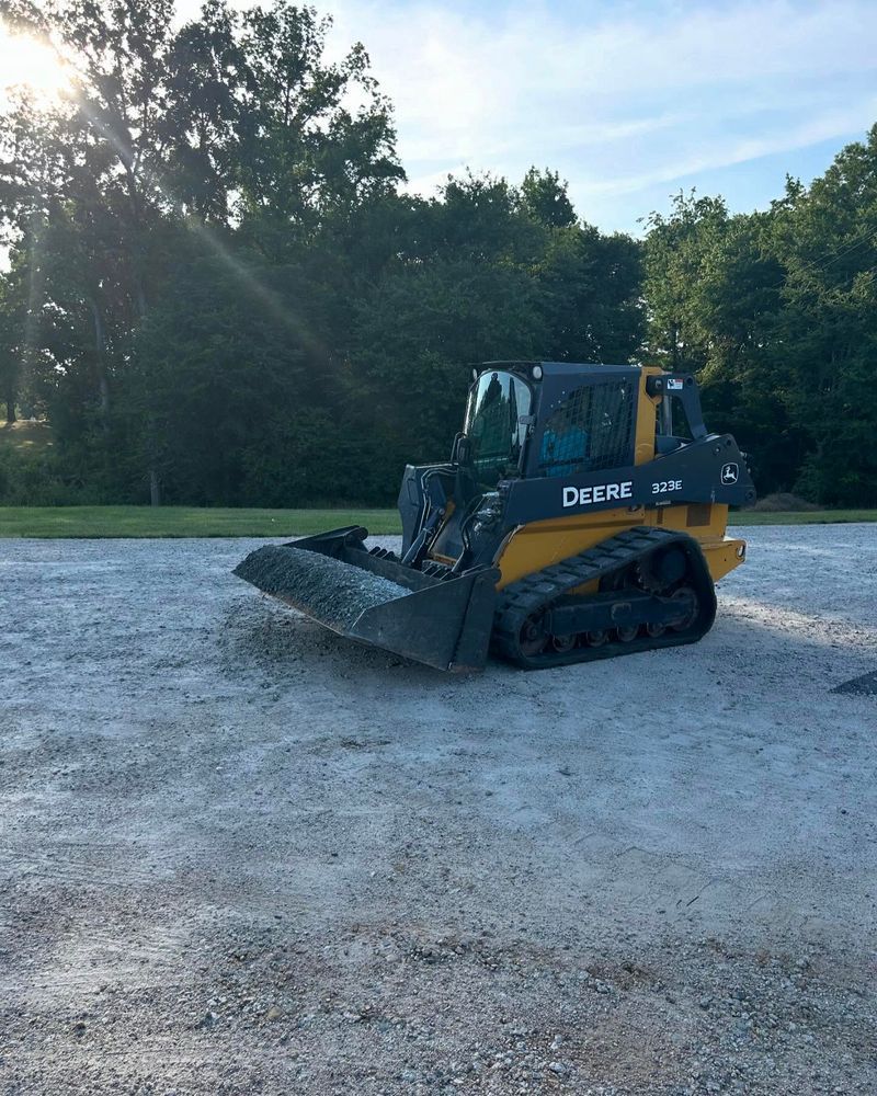 Yellow and black John Deere track skid steer loader on gravel, in front of trees.