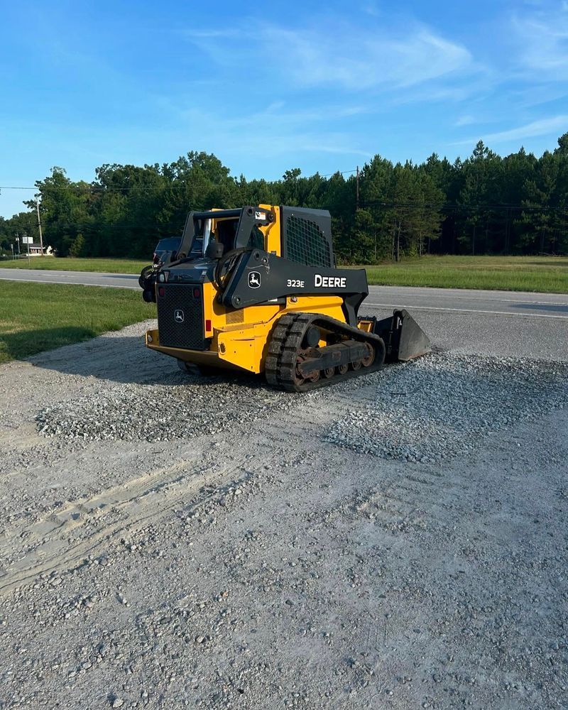 Yellow and black skid steer on gravel, parked on asphalt; trees and blue sky in background.