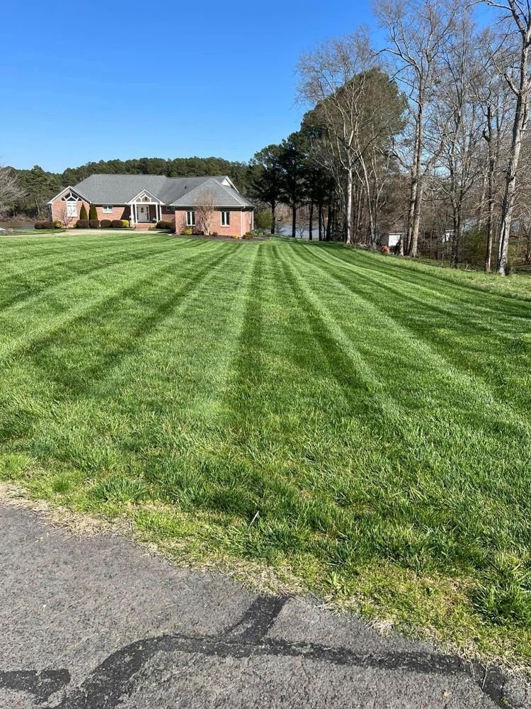 Lawn with a striped mowing pattern in front of a brick house. Trees and a body of water are visible in the background.