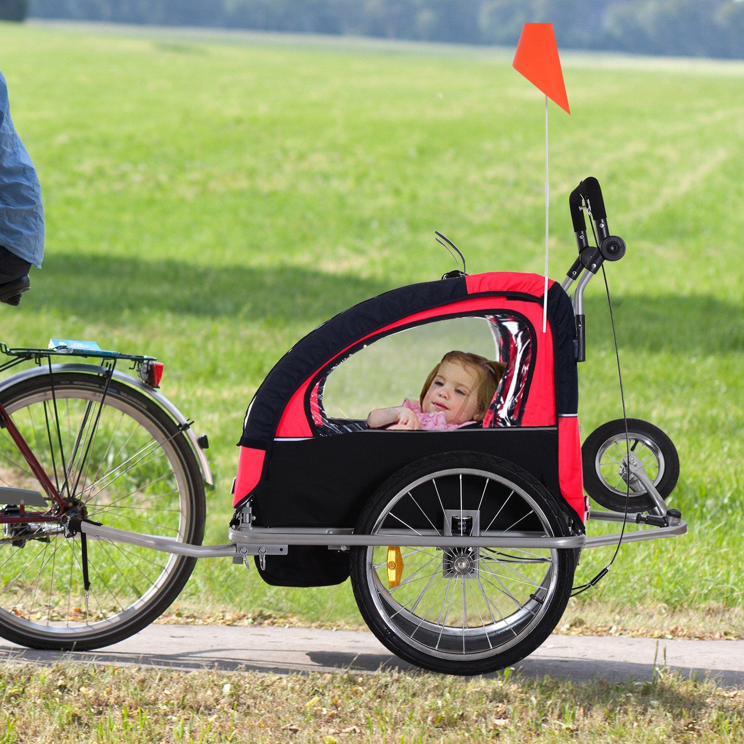 A child is sitting in a red and black trailer attached to a bicycle.