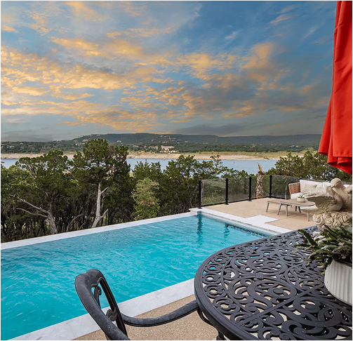 Infinity pool overlooking a lake and trees with a sunset sky. A table and chairs are in the foreground.