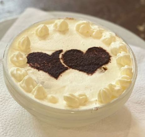 Dessert tiramisù in una ciotola trasparente, guarnito con panna, cuori di cacao e vortici di panna.