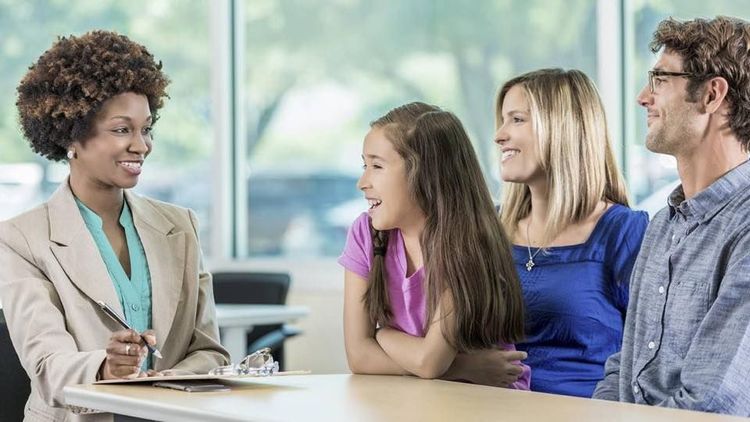 Woman in beige blazer speaks to a family: girl, mother, and father seated at a table, smiling.