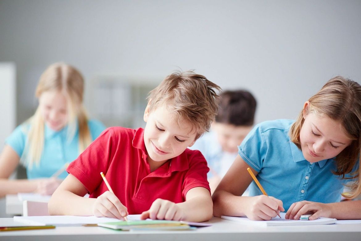 Children in a classroom writing at their desks; boy in red shirt smiles, girl in blue shirt focused.