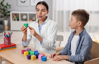 A woman in a lab coat shows a green block to a boy at a table with colorful blocks in a therapy setting.
