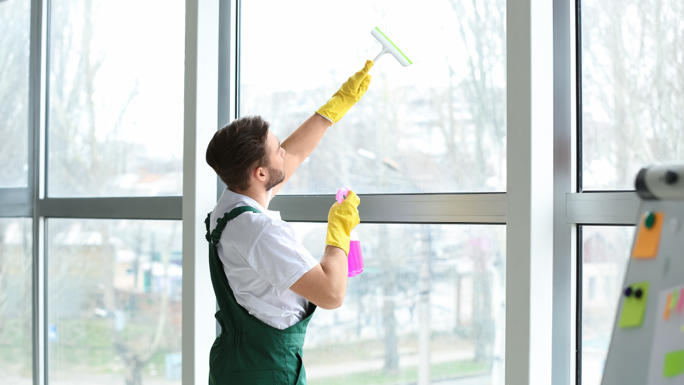 A worker in green overalls and yellow gloves uses a squeegee to clean a large office window.