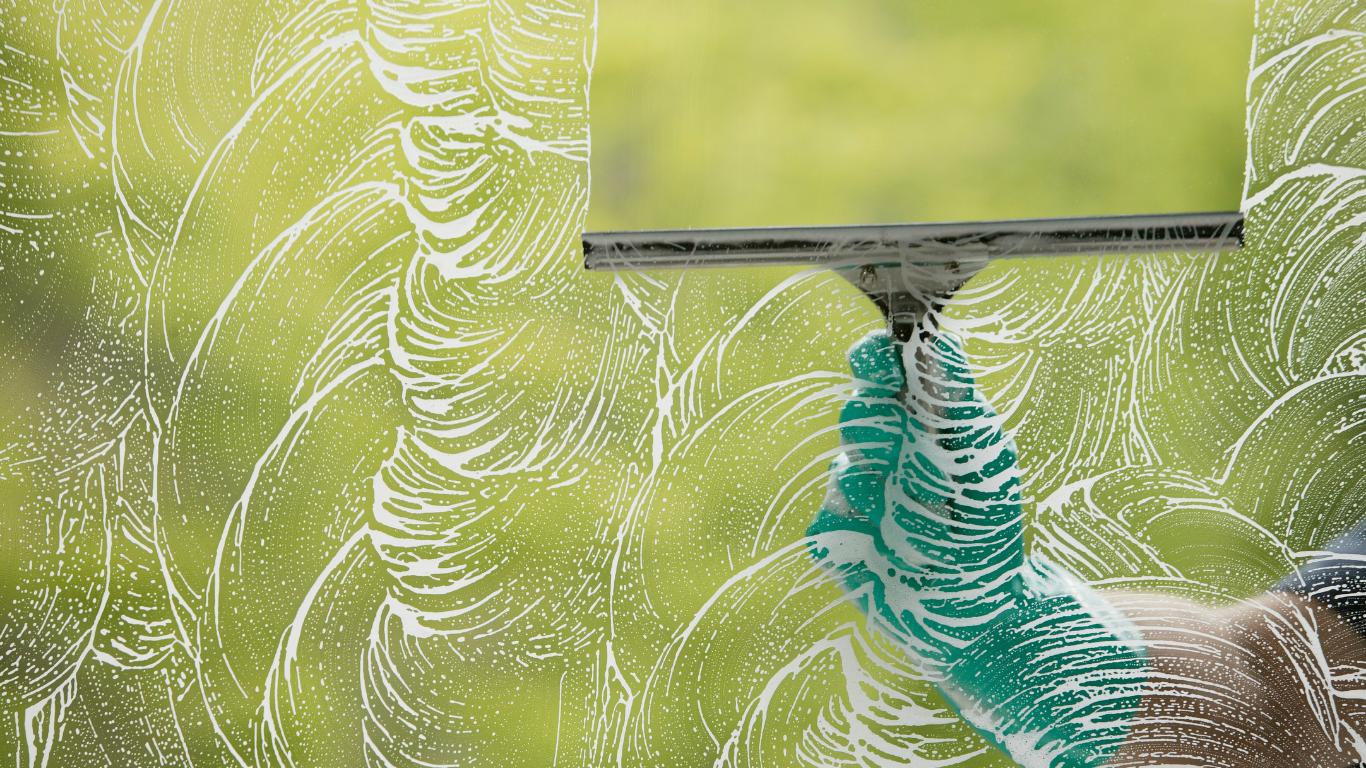A hand in a green patterned glove uses a squeegee to wipe soapy streaks off a glass window, revealing a clear view.