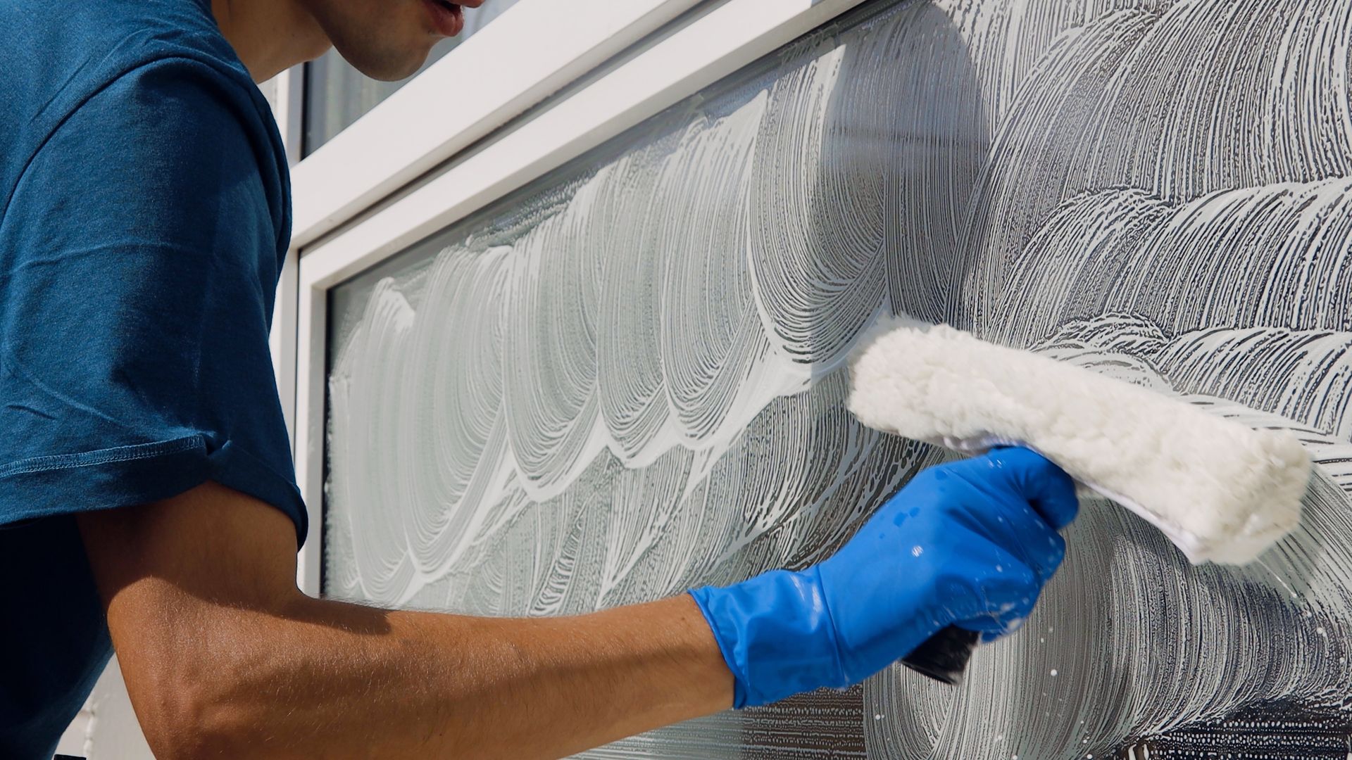 A person wearing a blue shirt and blue glove cleans a window with a soapy scrubber.