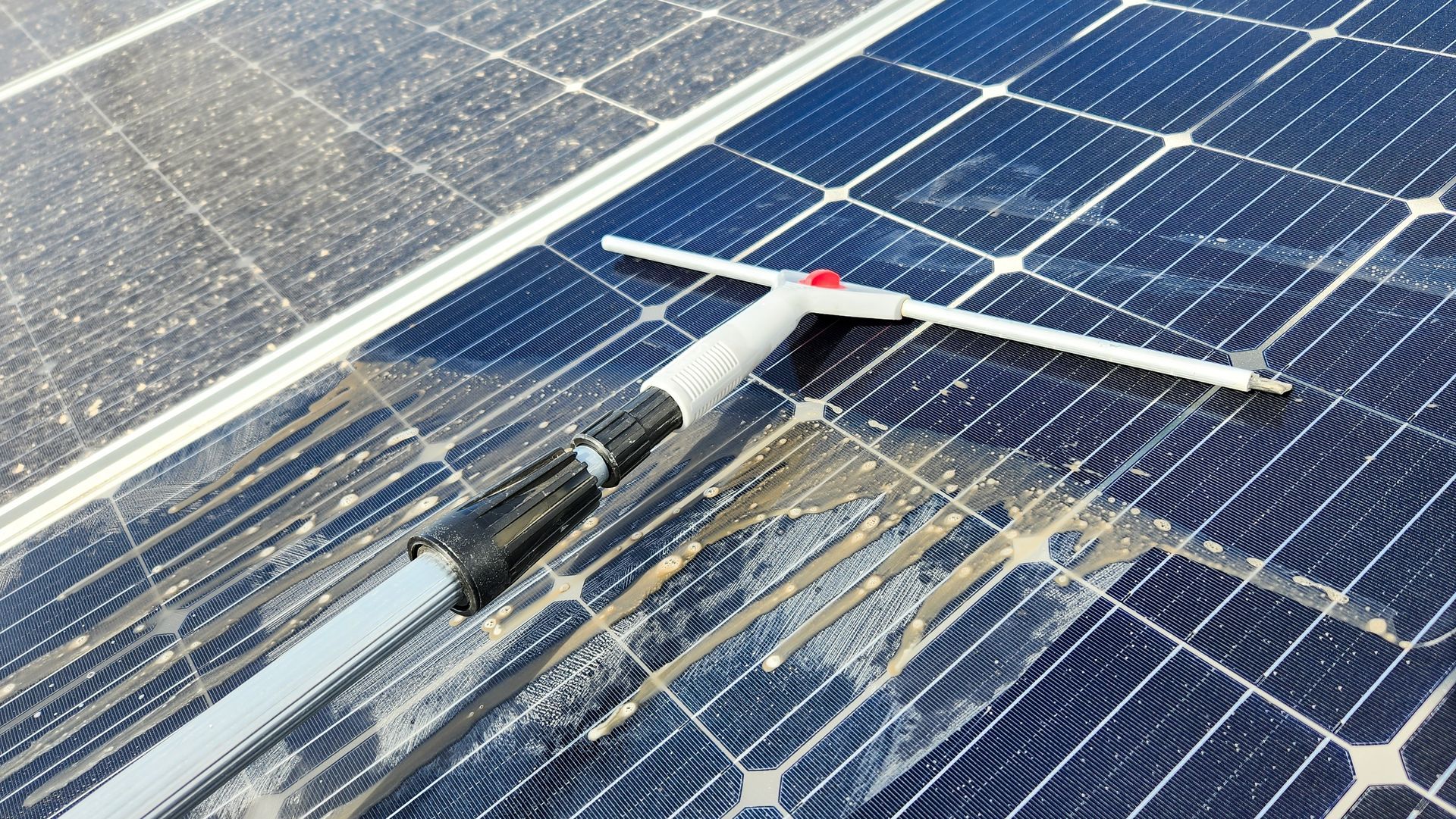 A long-handled squeegee cleans dirt and water from the surface of a blue-grid solar panel.