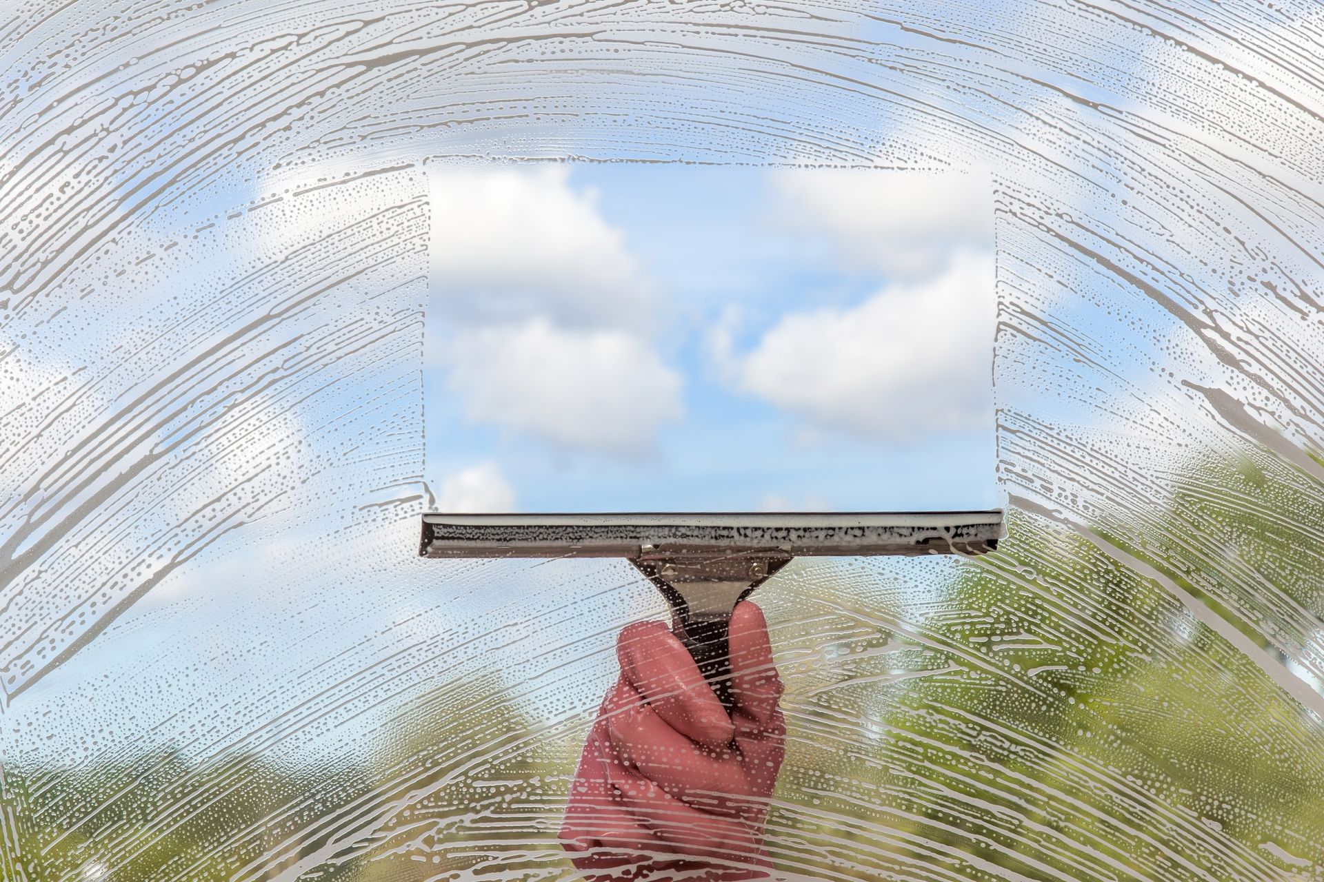 A gloved hand uses a squeegee to clear a soapy window, revealing a patch of blue sky and clouds behind it.